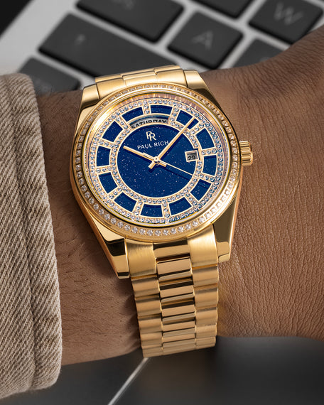 Gold watch with a blue aventurine dial, pavé diamond bezel, and day–date display shown on a wrist with a matching gold bracelet. product_visual, [lifestyle]