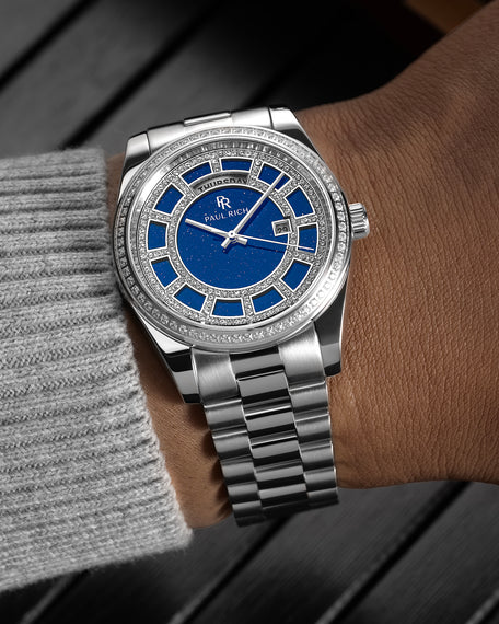 Silver watch with a blue aventurine dial, pavé diamond bezel, and day–date display shown on a wrist with a polished steel bracelet. product_visual, [lifestyle]