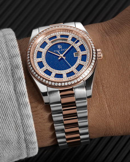 Rose gold and silver two-tone watch with a blue aventurine dial, pavé bezel, and day–date display shown on a wrist with a brushed metal bracelet. product_visual, [lifestyle]