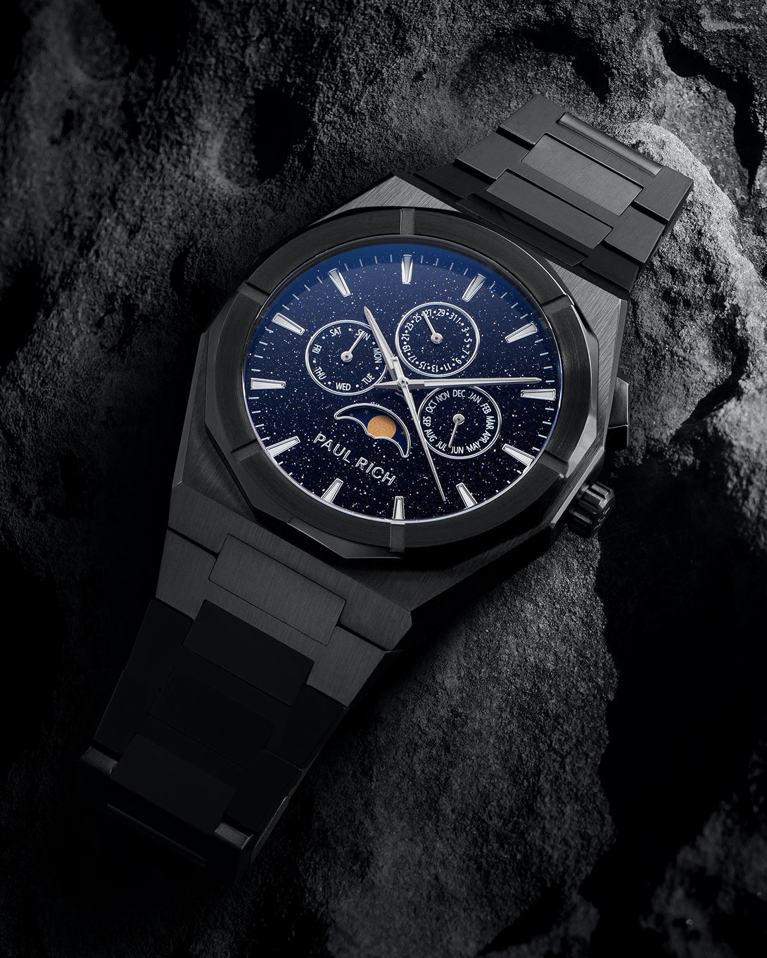Black steel bracelet watch with speckled blue dial, moonphase at 6 and three calendar subdials; angled on dark textured rock. product_visual