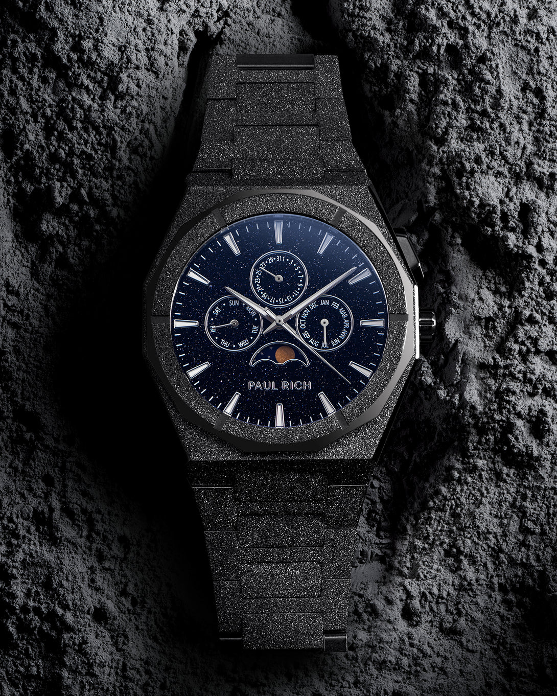 Front view of frosted black bracelet watch with speckled blue dial, moonphase at 6 and three calendar subdials; on gray textured rock. product_visual