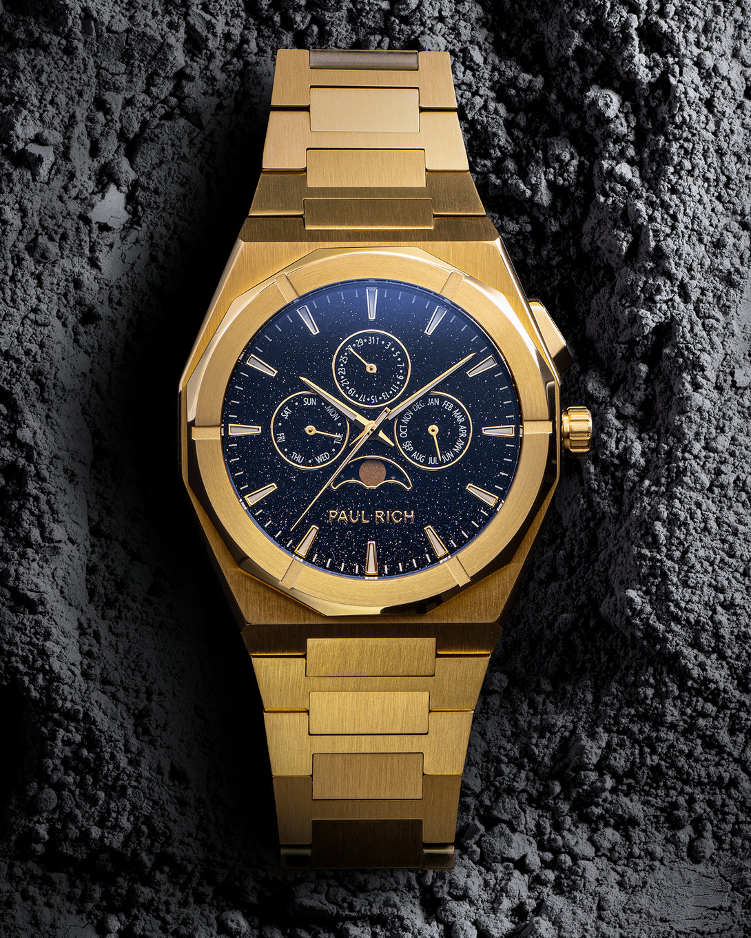 Front view of gold bracelet watch with speckled blue dial, moonphase at 6 and three calendar subdials; brushed links on gray textured rock. product_visual
