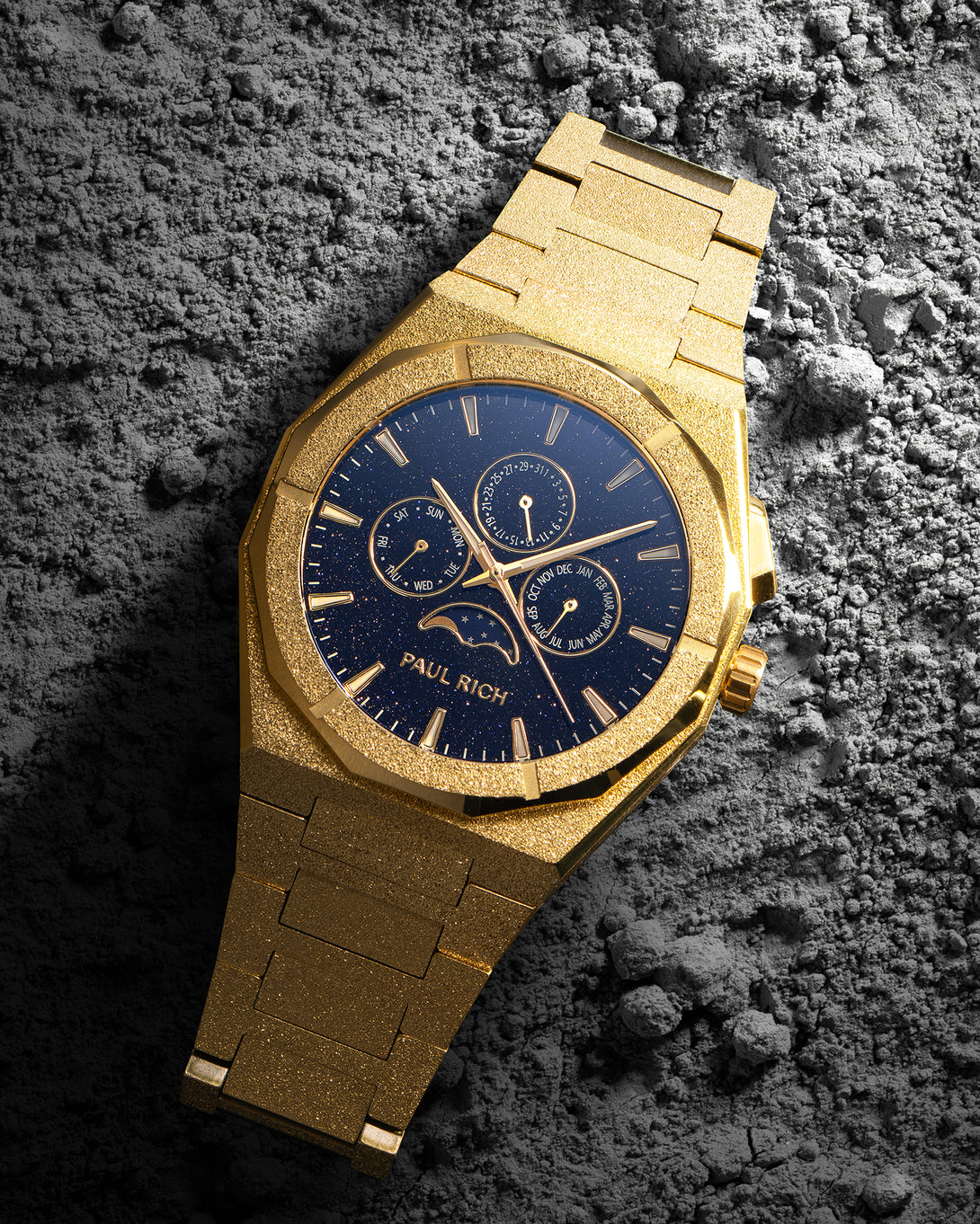 Frosted gold bracelet watch with speckled blue dial, moonphase at 6 and three calendar subdials; angled on gray textured rock. product_visual