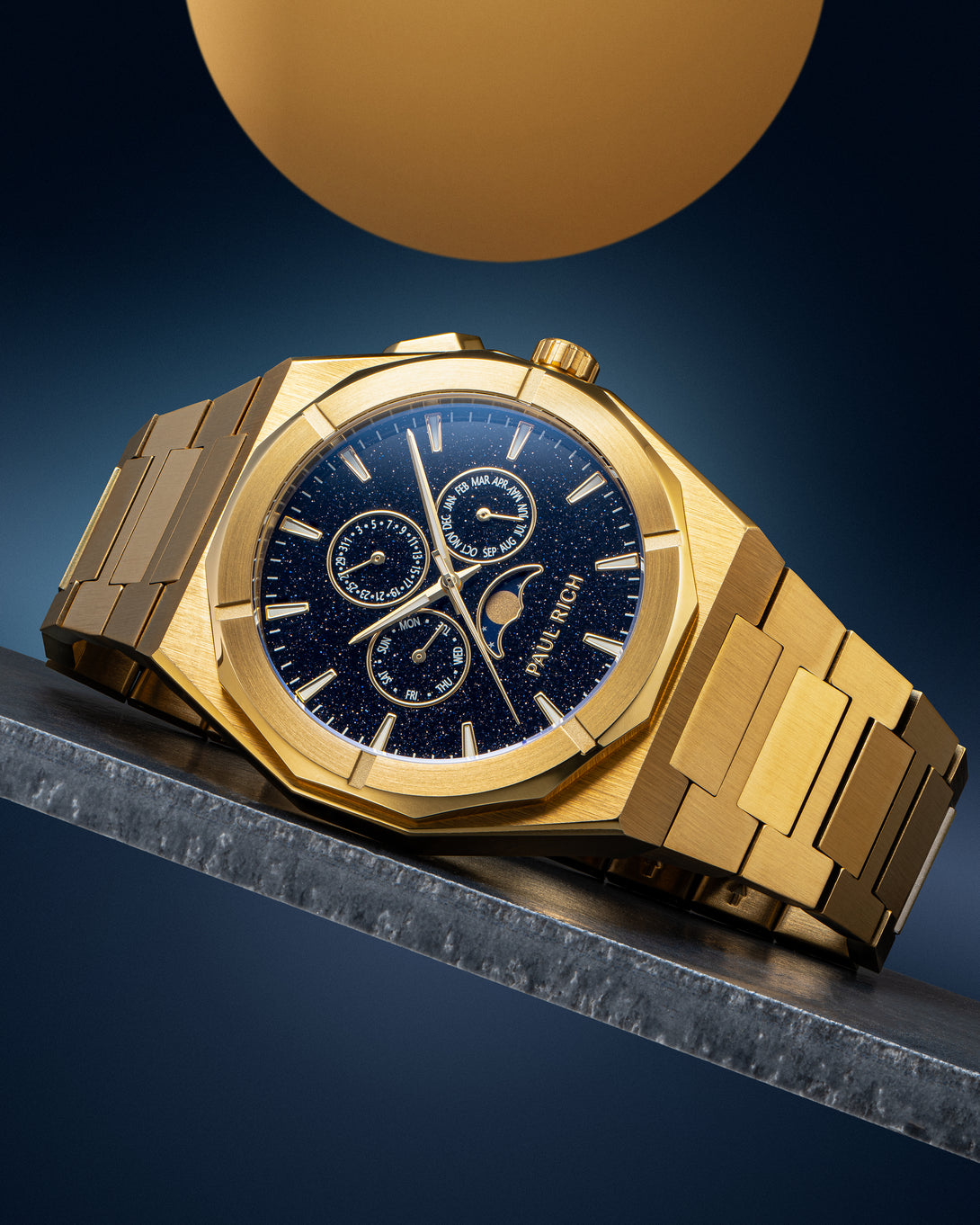 Gold-tone stainless steel watch with blue aventurine dial, moonphase, and subdials, displayed on a metal surface against a dark backdrop. product_visual