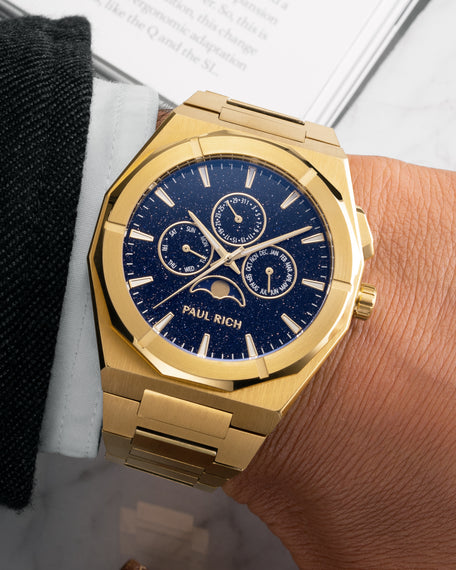 Brushed gold moonphase watch on wrist with deep blue aventurine dial, calendar subdials, and gold bracelet. [lifestyle], product_visual