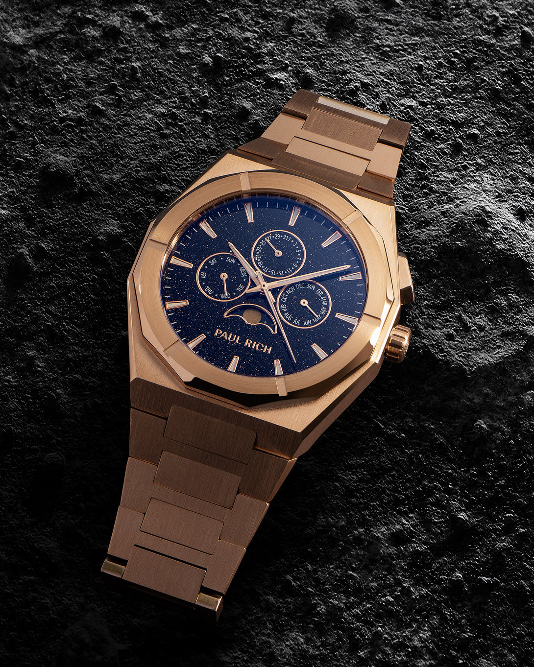 Paul Rich rose-gold bracelet watch with speckled blue dial, moonphase and calendar subdials; brushed links on dark textured stone. product_visual