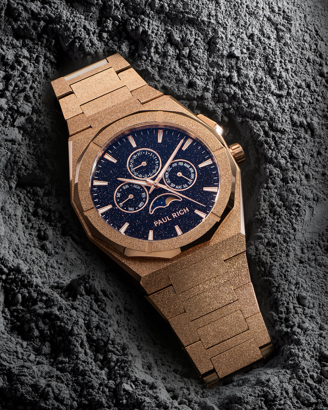 Frosted rose-gold bracelet watch with speckled blue dial, moonphase at 6 and three calendar subdials; angled on gray textured rock. product_visual