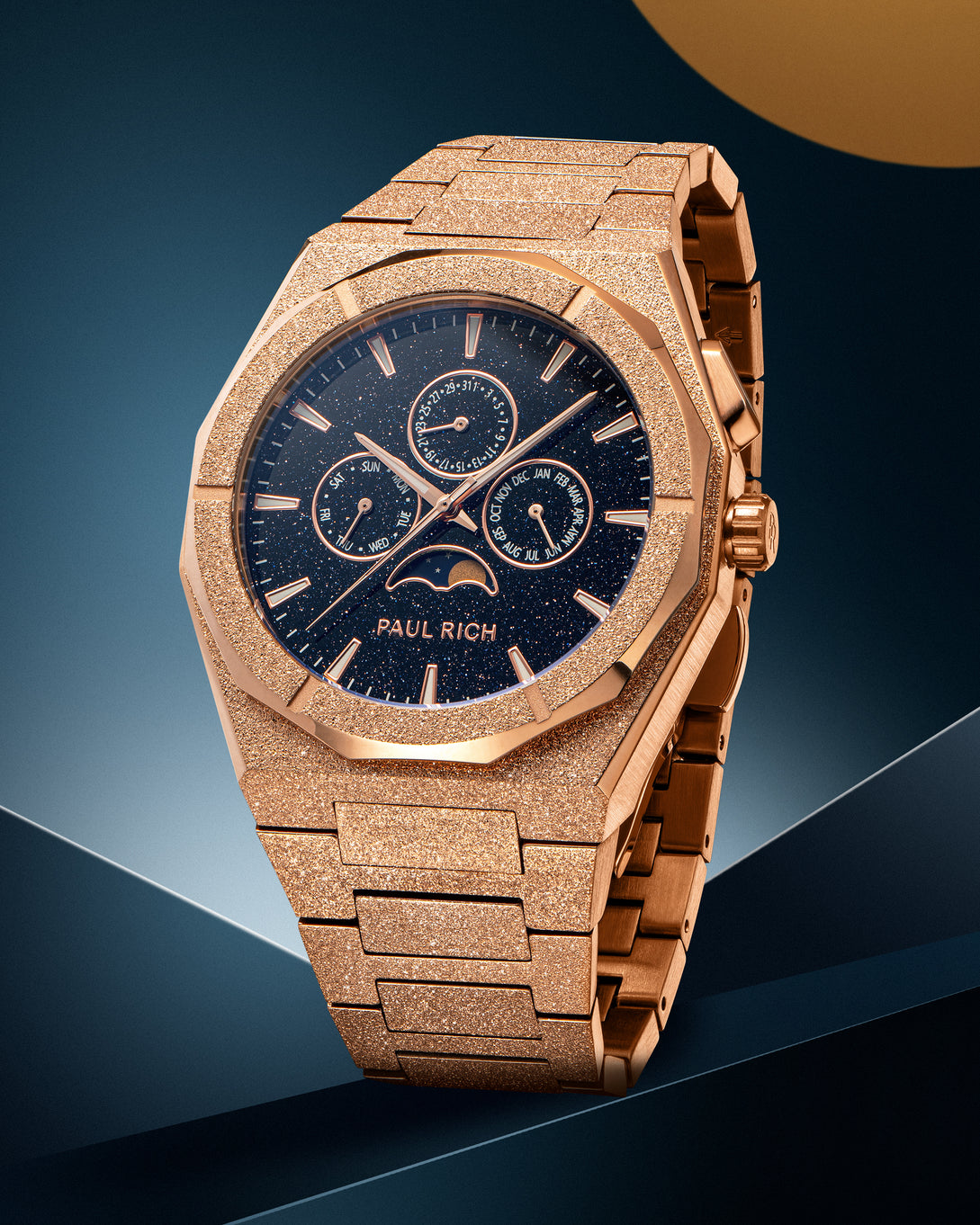 Frosted rose gold-tone stainless steel watch with blue aventurine dial, moonphase, and subdials, displayed on a geometric blue and gold backdrop. product_visual
