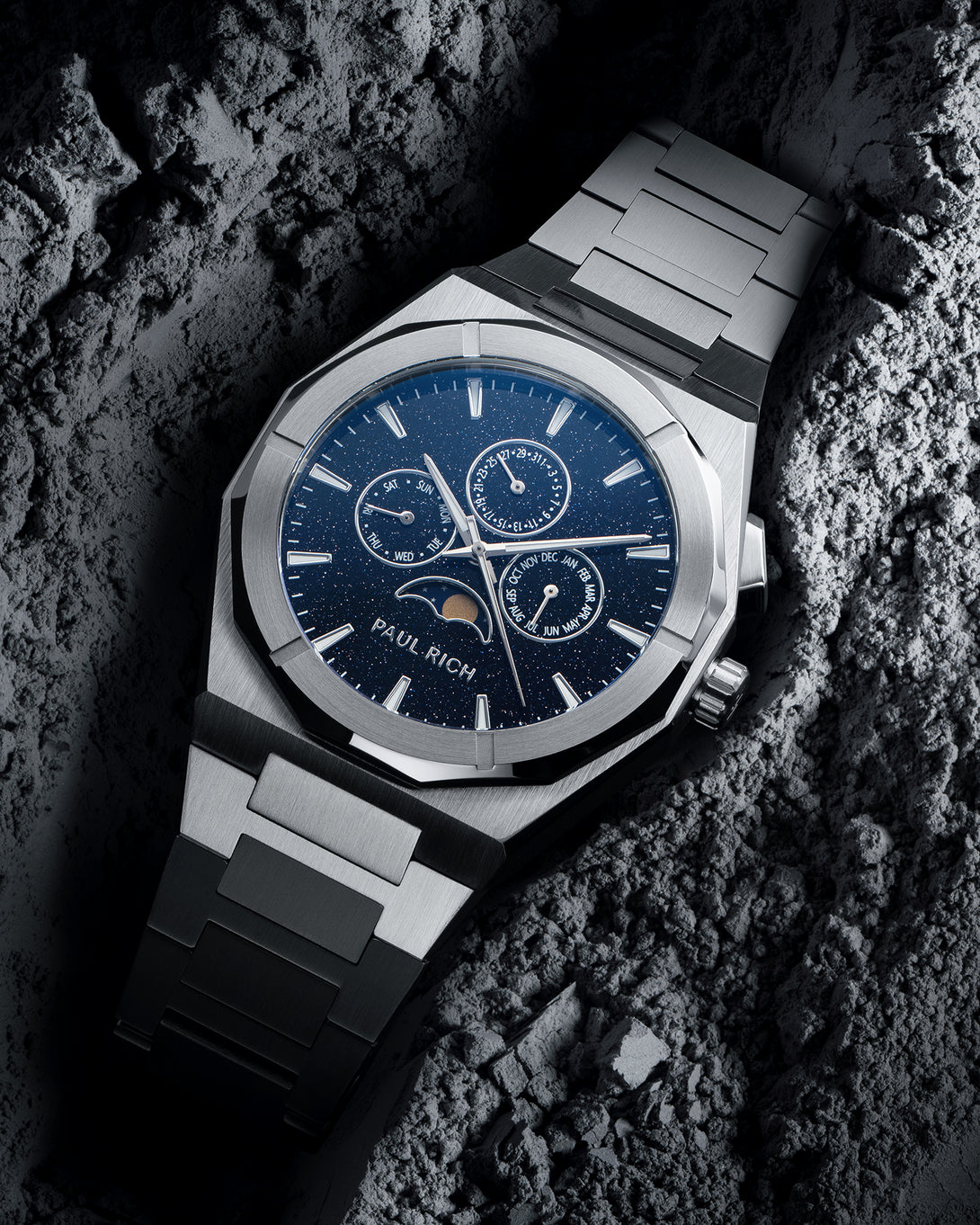 Silver steel bracelet watch with speckled blue dial, moonphase at 6 and three calendar subdials; angled on gray textured rock. product_visual