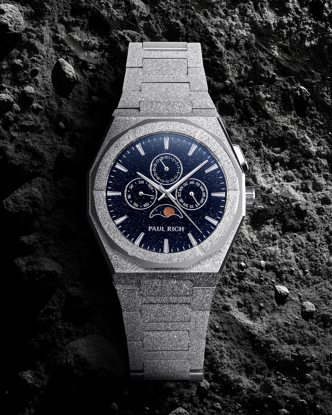 Frosted silver bracelet watch with deep blue speckled dial, moonphase at 6 and three calendar subdials, set on dark textured rock. product_visual