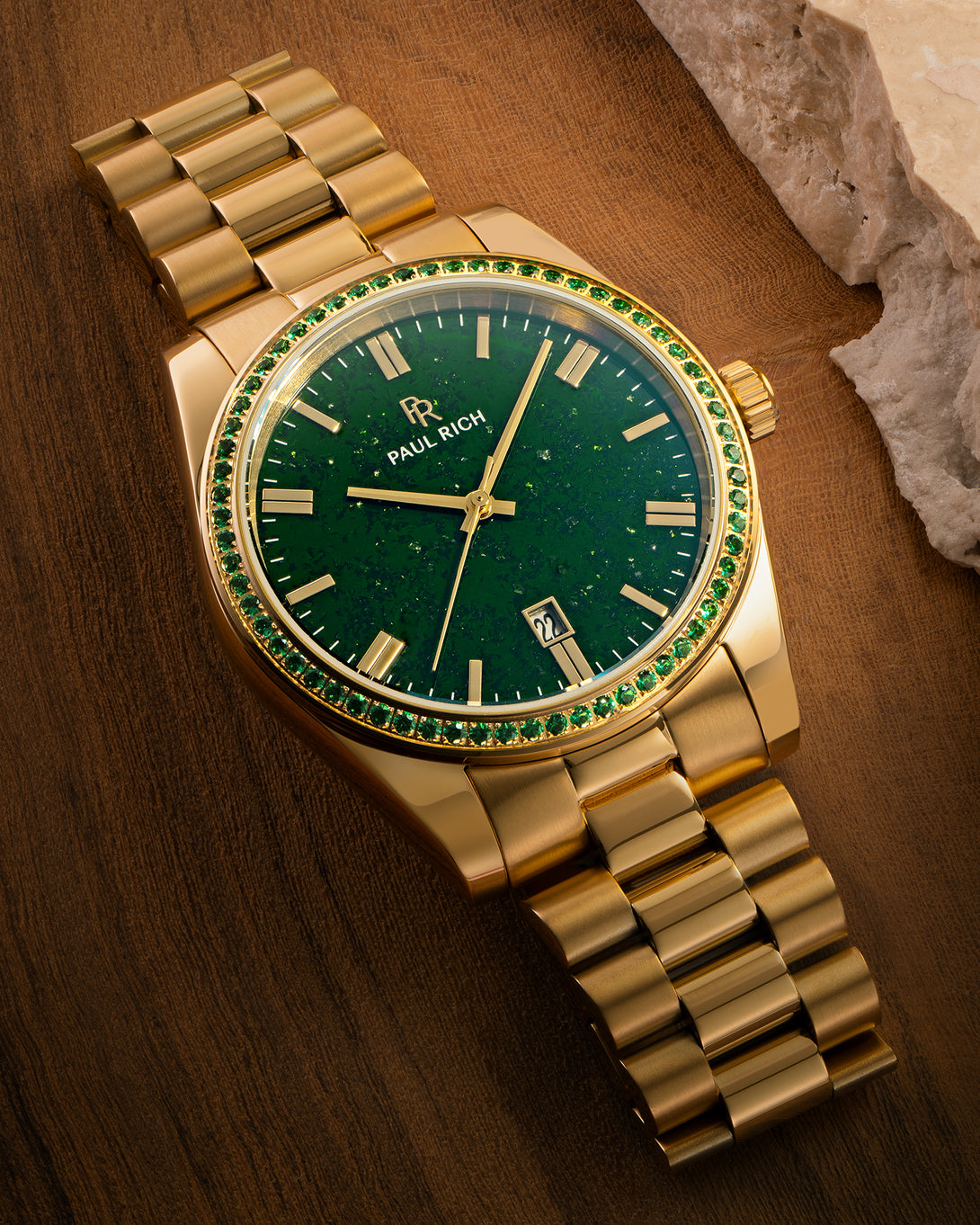Gold watch with green aventurine dial and green gemstone-set bezel displayed on a wooden surface near a stone edge, showing the Paul Rich logo and date window. product_visual