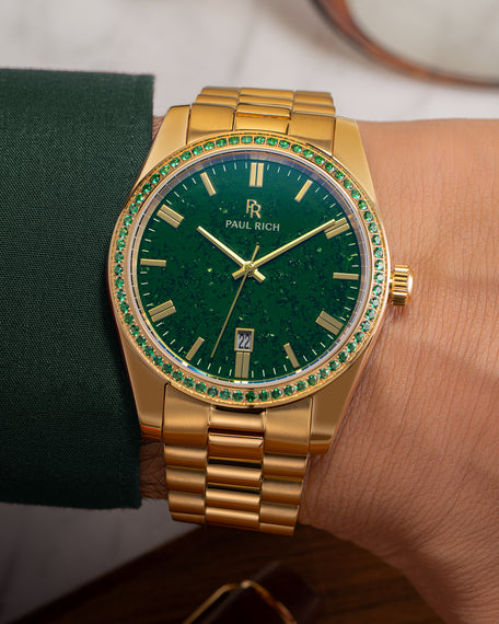 Gold watch with green aventurine dial and green gemstone-set bezel on a wrist wearing a dark green sleeve, showing the Paul Rich logo and date window. product_visual [lifestyle]