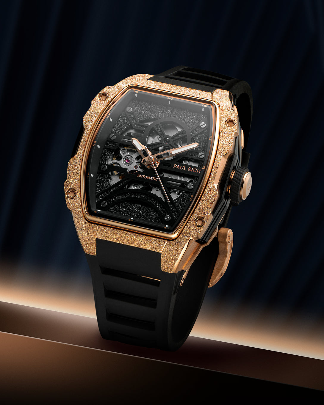 Rose gold frosted tonneau case watch with open skeleton dial and black strap on a dark blue draped background. product_visual