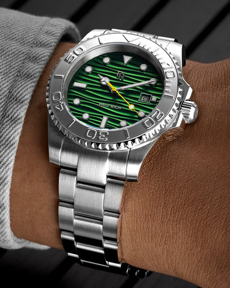 Stainless steel watch with a green wave-textured dial and yellow seconds hand shown on a wrist, highlighting the bezel, markers, and brushed bracelet. [lifestyle] product_visual