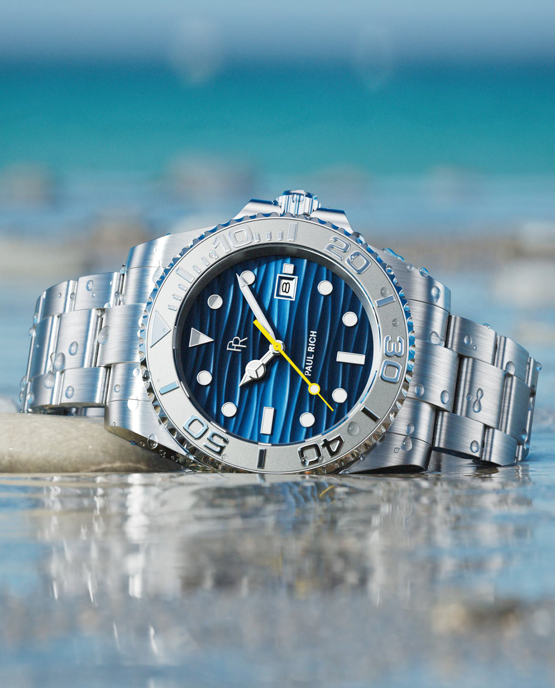 Silver dive watch with blue wave-textured dial, steel bracelet, rotating bezel, luminous markers, photographed in shallow water with ocean backdrop. product_visual
