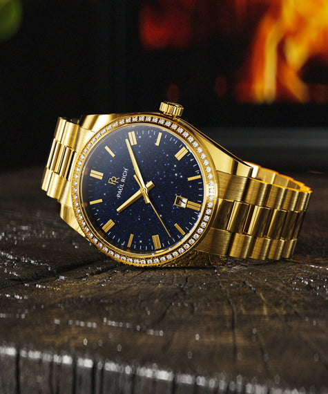 Gold Paul Rich watch with a blue aventurine dial and gem-set bezel, resting on wood with a fireplace glowing in the background. [lifestyle] product_visual