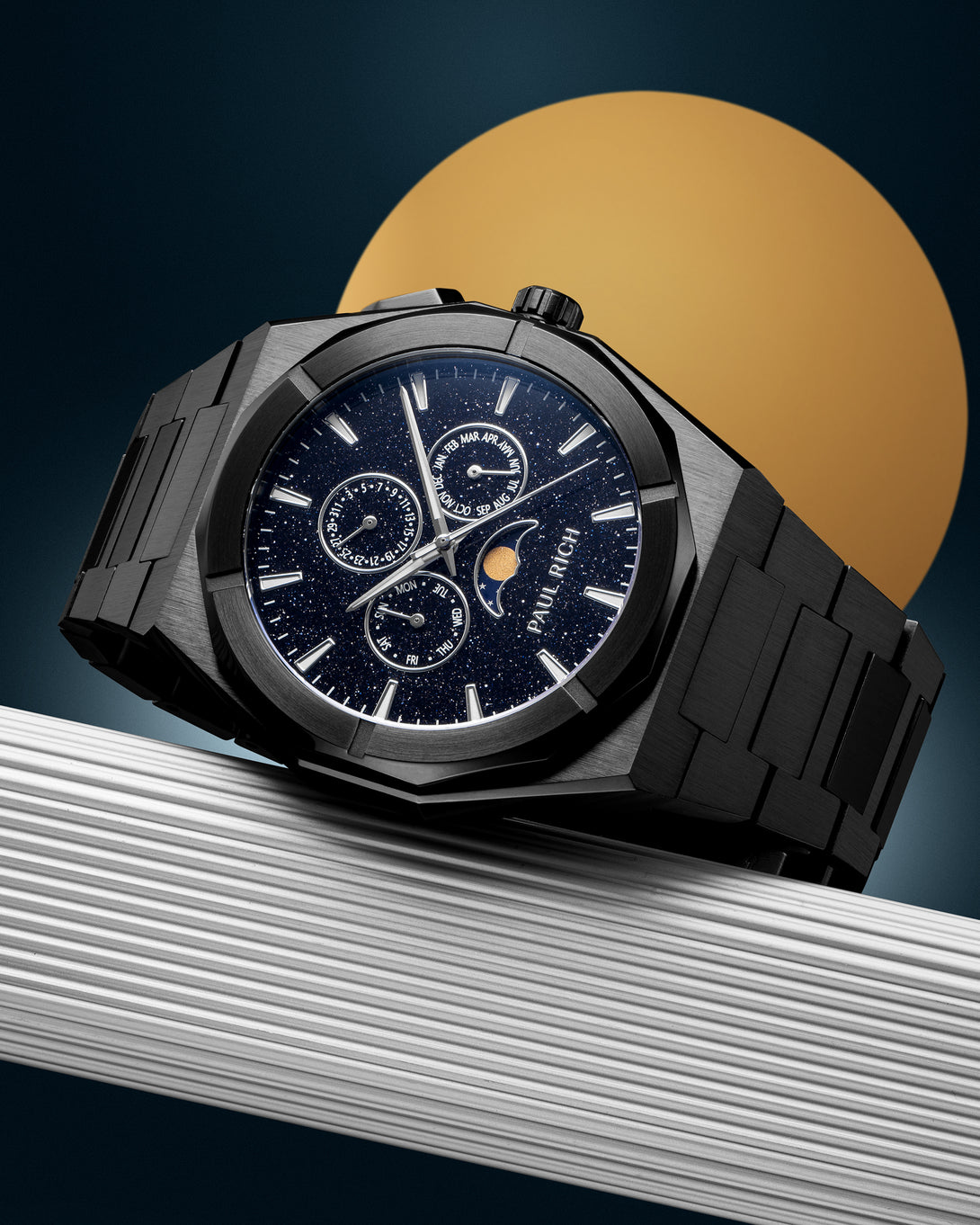 Black stainless steel watch with blue aventurine dial, moonphase, and subdials, displayed on a ribbed silver surface against a gold circle backdrop. product_visual